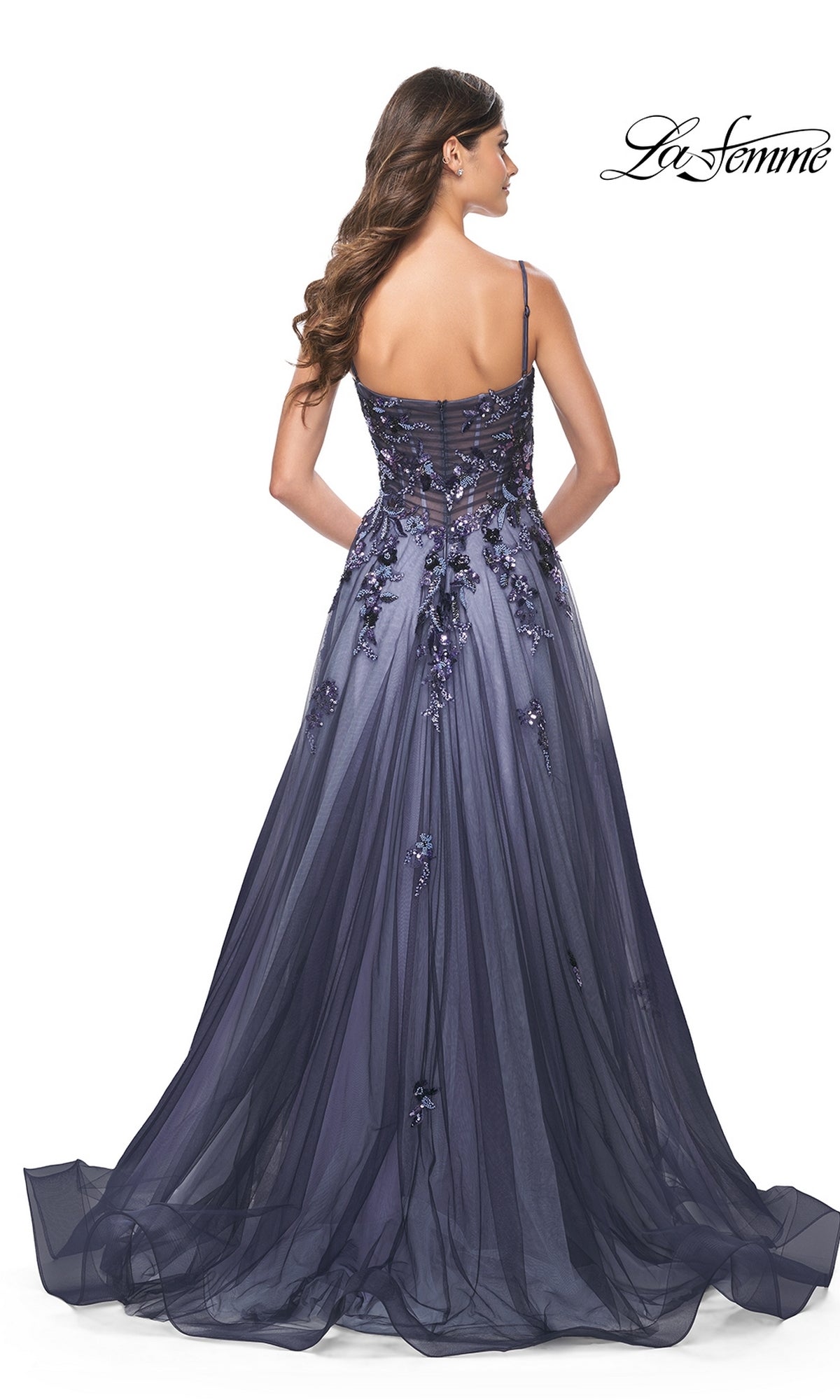 A woman with long brown hair stands with her back to the camera, wearing the La Femme Long Navy Blue A-Line Prom Dress 32185—a floor-length gown with sequin-lace floral appliqué and a sheer back.